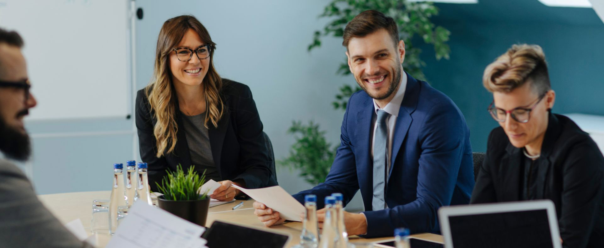4 people sitting at a table signing legal documents in an office, some wearing suits, smiling and collaborating on a business contract.